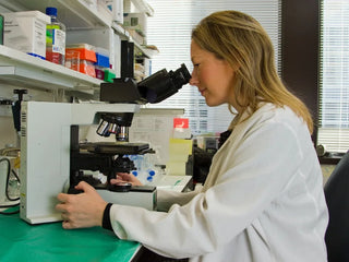 A woman in a white lab coat looks through a microscope in a laboratory setting with shelves and equipment in the background.