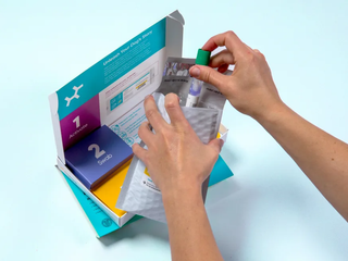 A hand placing a capped Embark DNA swab collection tube into a grey return mailer next to the opened product box.
